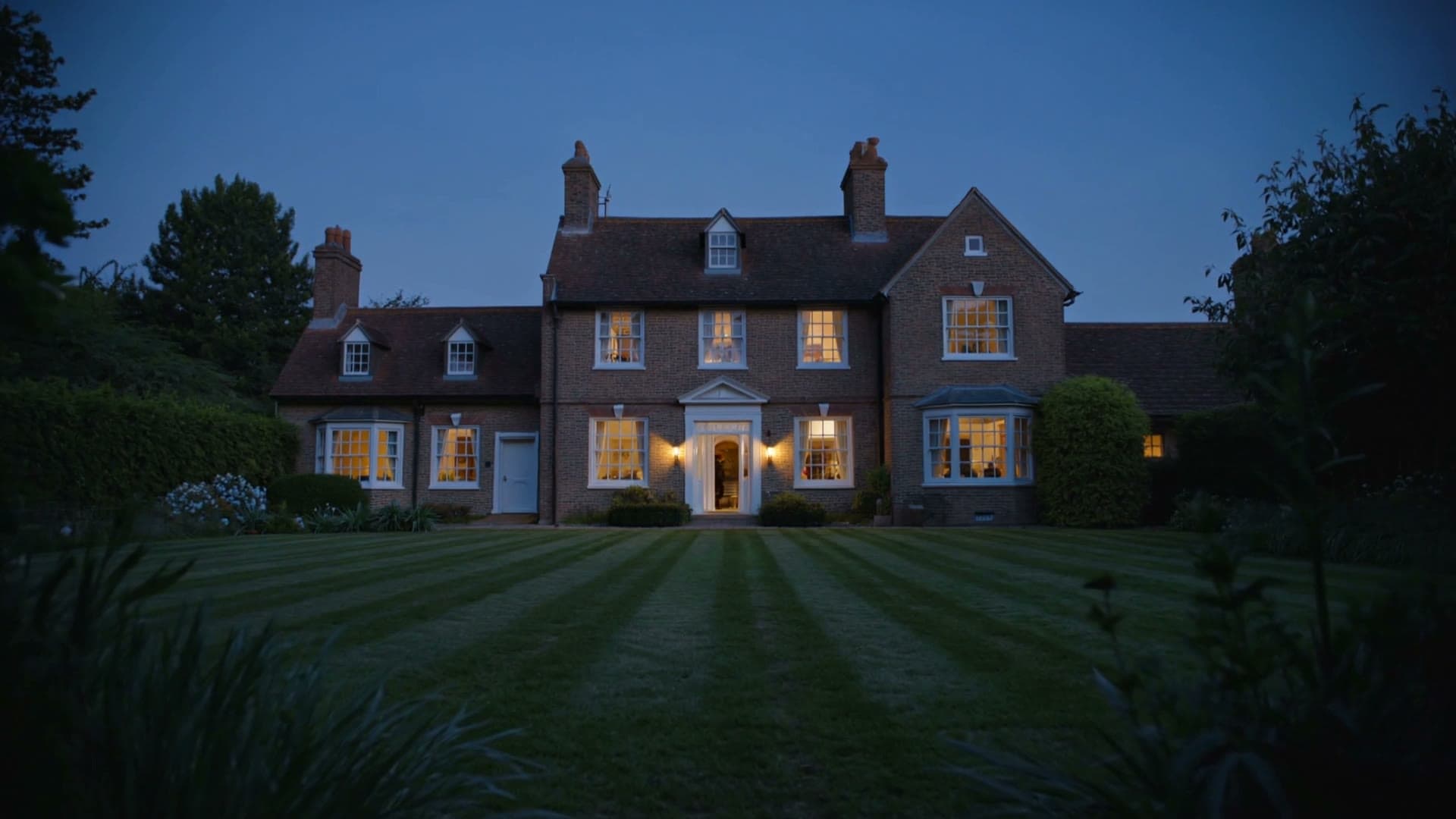 A period country house at dusk, its windows lit warm against a blue evening sky